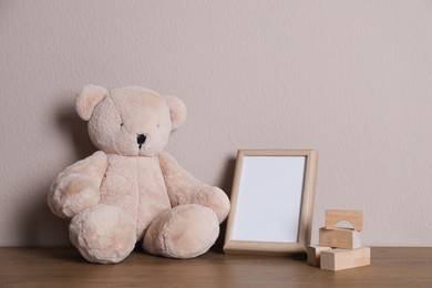 Photo of Empty photo frame, teddy bear and building blocks on wooden table near grey wall. Space for design