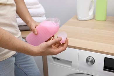Photo of Man pouring fabric softener from bottle into cap near washing machine indoors, closeup