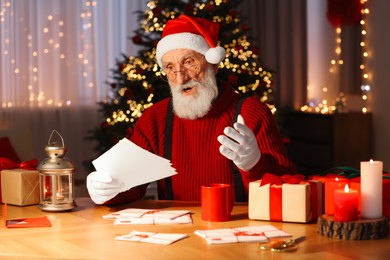 Santa Claus reading letter at his workplace in room with Christmas tree