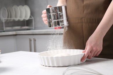 Photo of Woman sieving flour into baking dish at table in kitchen, closeup