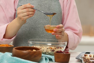 Making granola. Woman adding honey into bowl with mixture of oat flakes and other ingredients at table in kitchen, closeup