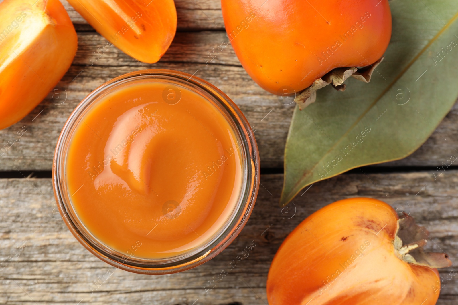 Photo of Delicious persimmon jam in glass jar and fresh fruits on wooden table, flat lay