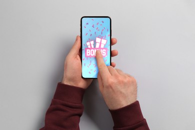 Image of Bonus gaining. Man using smartphone on light grey background, top view. Illustration of open gift boxes, word and falling confetti on device screen