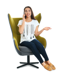 Young woman sitting in armchair and talking by smartphone on white background