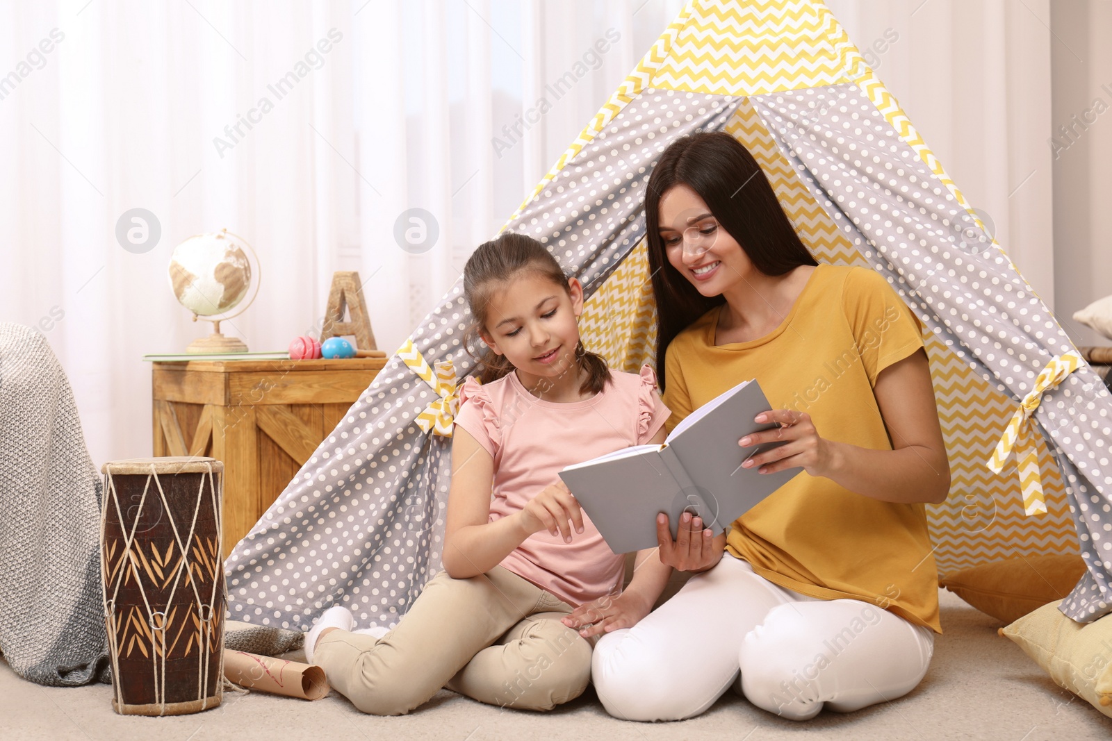 Photo of Mother and daughter reading book near toy wigwam at home