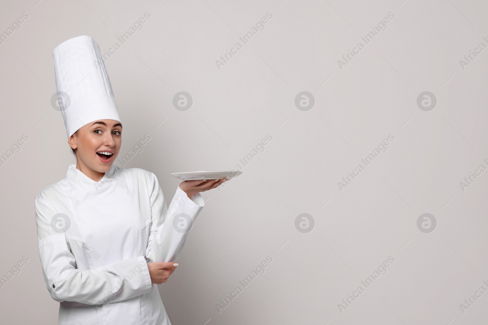 Photo of Happy professional confectioner in uniform holding empty plate on light grey background. Space for text