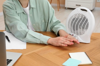 Photo of Young woman warming hands near modern electric fan heater at wooden table indoors, closeup