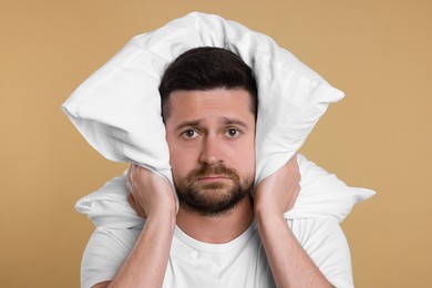 Photo of Unhappy man covering ears with pillow on beige background. Insomnia problem
