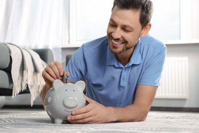 Photo of Happy man putting money into piggy bank on floor indoors