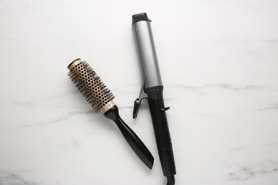 Curling iron and round hair brush on white marble background, flat lay