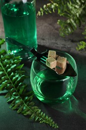 Photo of Absinthe in glass, spoon with brown sugar cubes and fern leaves on table, closeup. Alcoholic drink