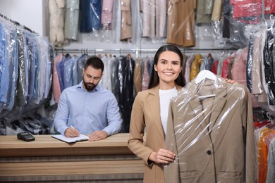 Photo of Dry-cleaning service. Happy woman holding hanger with coat indoors. Worker taking notes at workplace