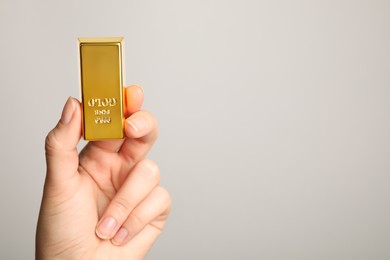 Photo of Woman holding shiny gold bar on light grey background, closeup. Space for text