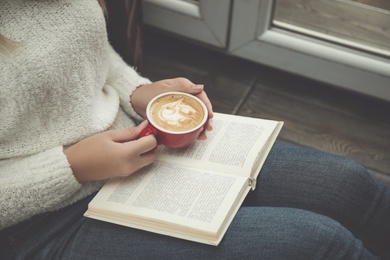 Woman with cup of coffee reading book near window indoors, closeup