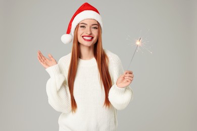 Photo of Young woman in Santa hat with burning sparkler on light grey background. Christmas celebration