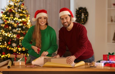 Photo of Happy couple in Santa hats decorating Christmas gift with wrapping paper at table in room