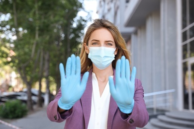 Photo of Woman in protective face mask showing stop gesture outdoors. Prevent spreading of coronavirus