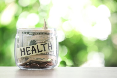 Photo of Glass jar with money and label HEALTH on table against blurred background. Space for text