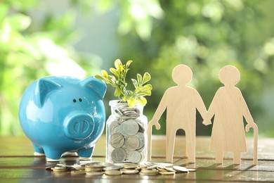 Pension savings. Figure of senior couple, piggy bank, coins and green twig on wooden table outdoors