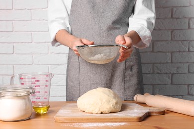 Woman sprinkling flour over dough at table near white brick wall, closeup