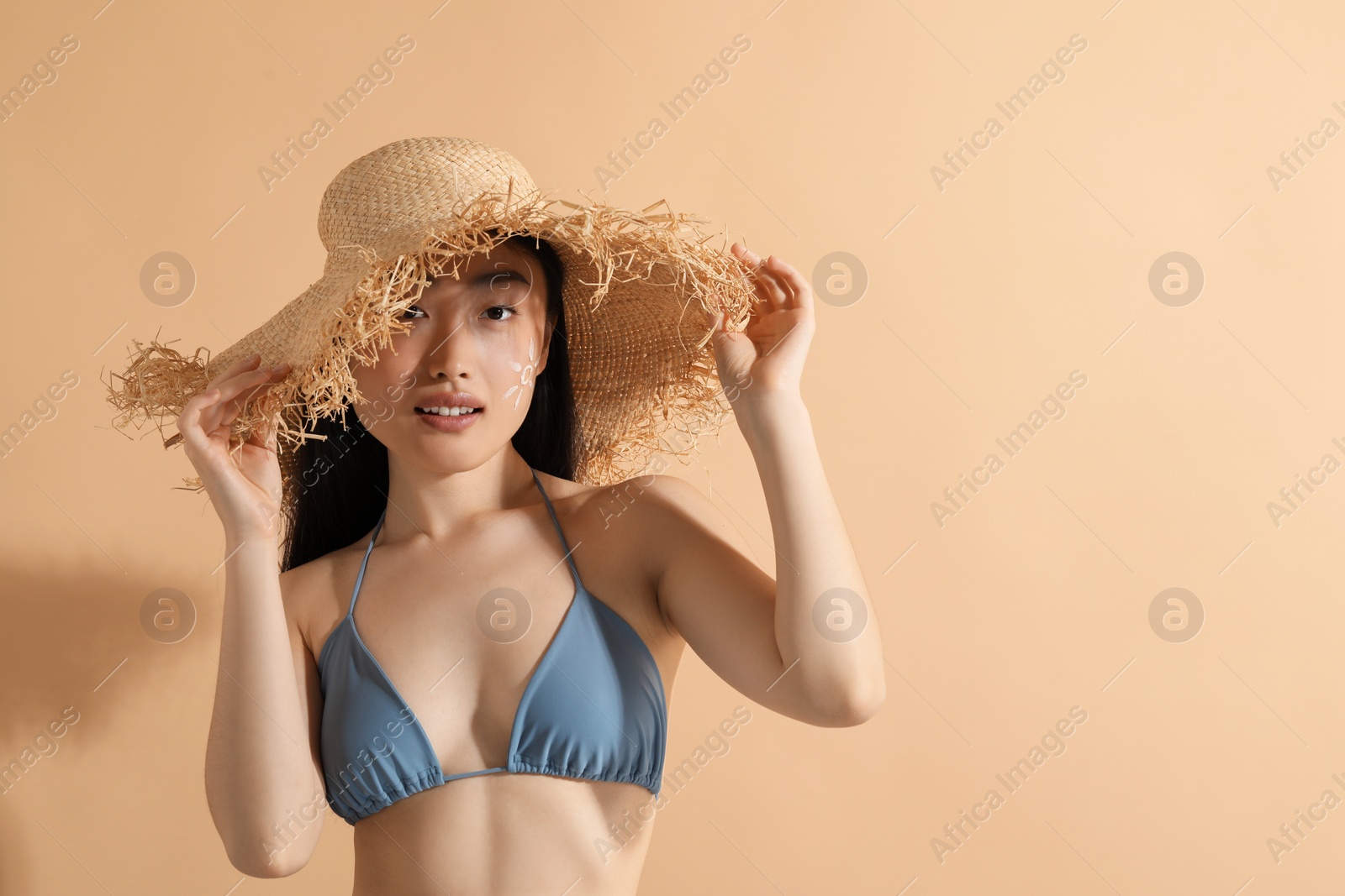 Photo of Beautiful young woman in straw hat with sun protection cream on her face against beige background, space for text