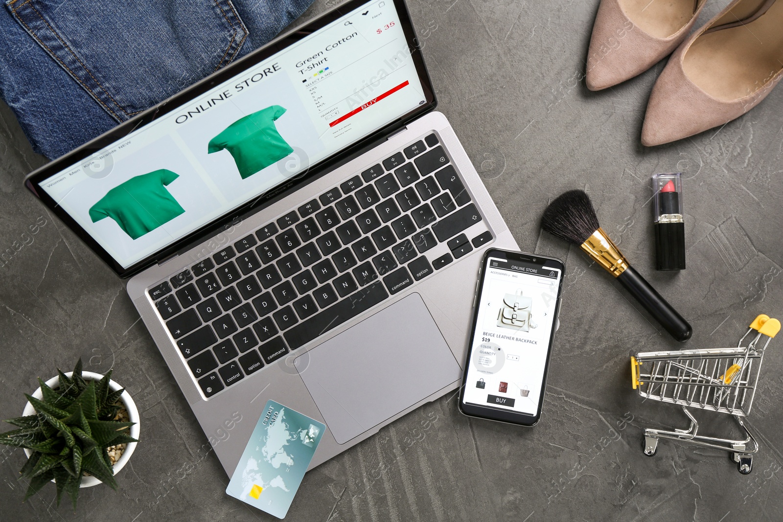 Photo of Online store website on laptop screen. Computer, smartphone, shopping cart, credit card, accessories and houseplant on grey table, flat lay