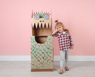 Photo of Little girl and cardboard costume of dinosaur near color wall indoors. Space for text