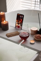 Photo of White wooden tray with smartphone, glass of wine and burning candles on bathtub in bathroom