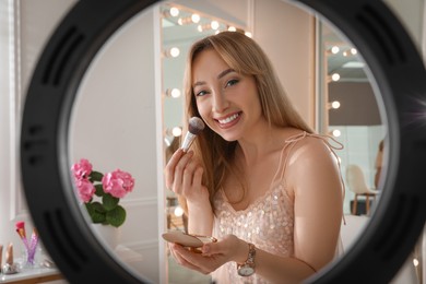 Beauty blogger with makeup brush and powder recording video in dressing room at home, view through ring lamp