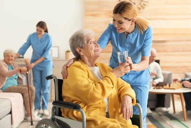 Photo of Nurse giving glass of water to elderly woman in wheelchair at retirement home. Assisting senior people