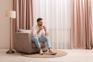 Photo of Smiling man drinking tea while resting on armchair near window with beautiful curtains at home. Space for text