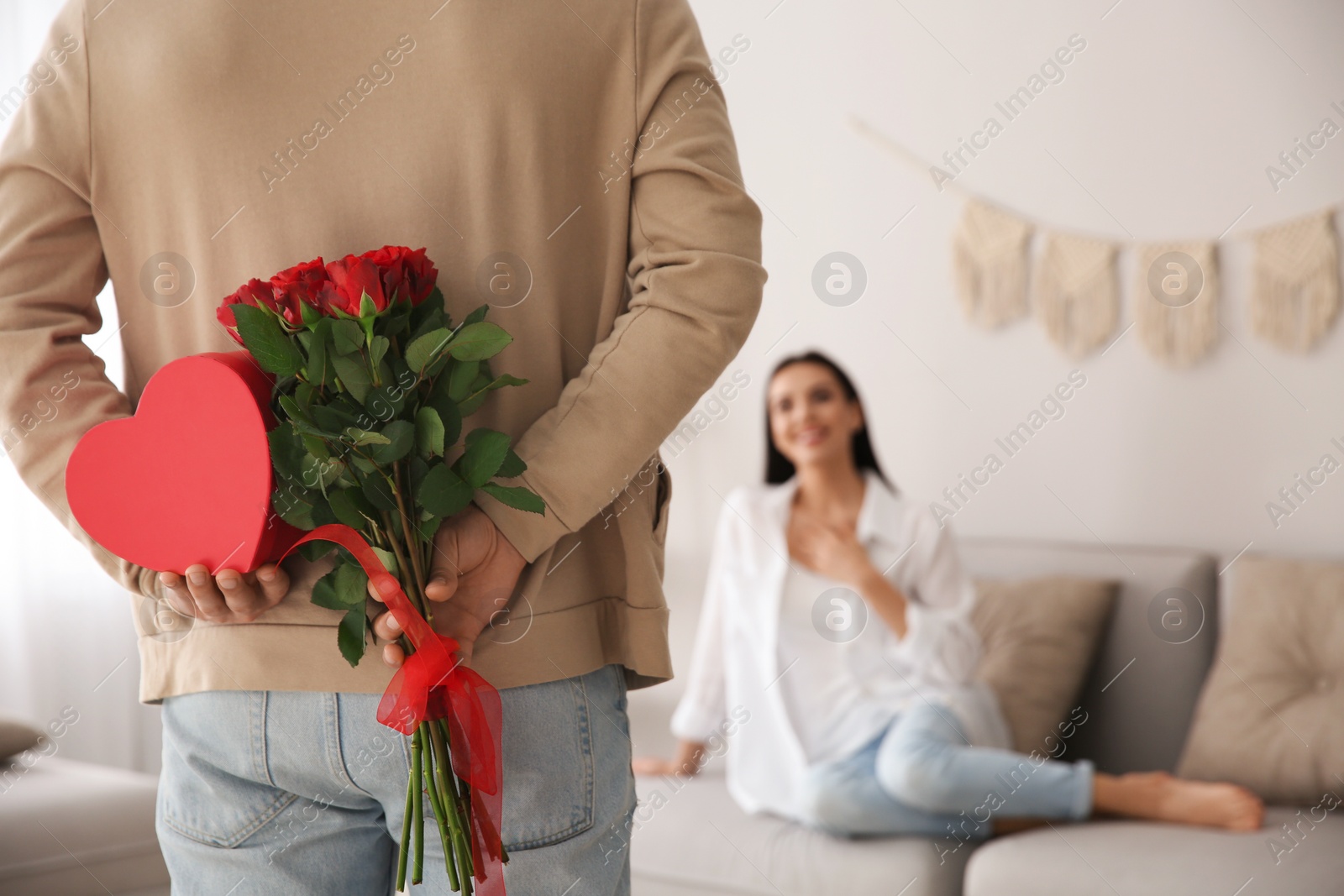 Photo of Man hiding beautiful bouquet and gift box behind his back at home, closeup. Valentine's day celebration