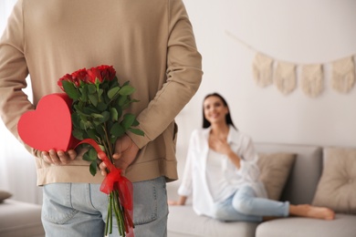 Man hiding beautiful bouquet and gift box behind his back at home, closeup. Valentine's day celebration