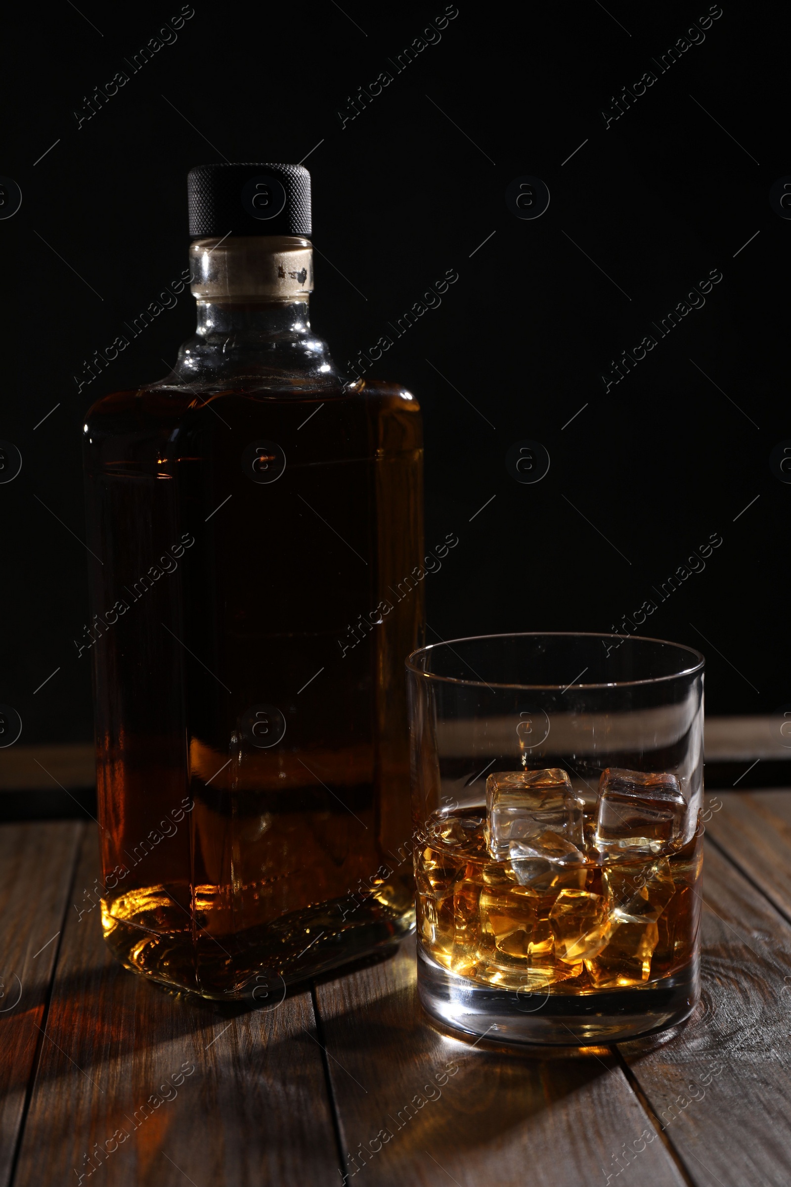 Photo of Whiskey with ice cubes in glass and bottle on wooden crate against black background