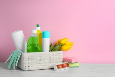 Plastic basket with different cleaning supplies and beautiful spring flowers on white wooden table against light pink background. Space for text