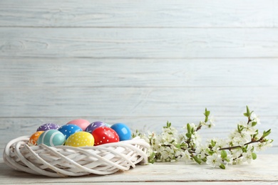 Photo of Painted Easter eggs in nest and blossoming branches on table. Space for text