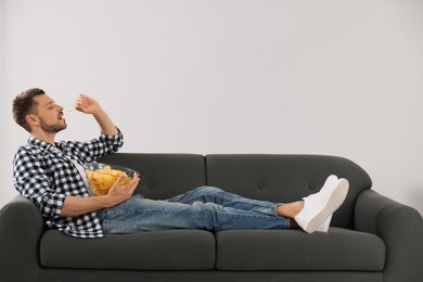 Handsome man eating potato chips on sofa near light wall
