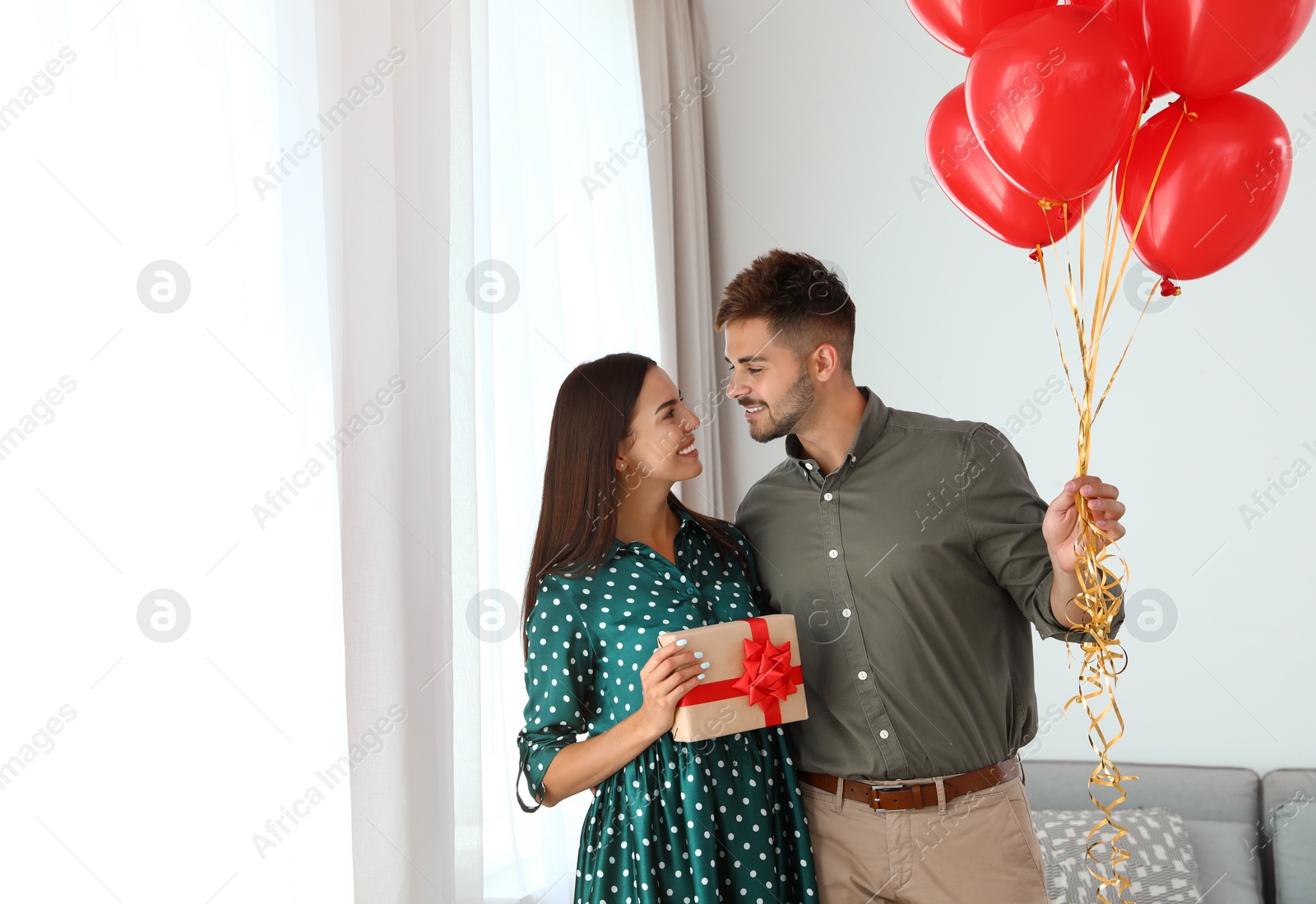 Photo of Young couple with air balloons and gift box at home. Celebration of Saint Valentine's Day