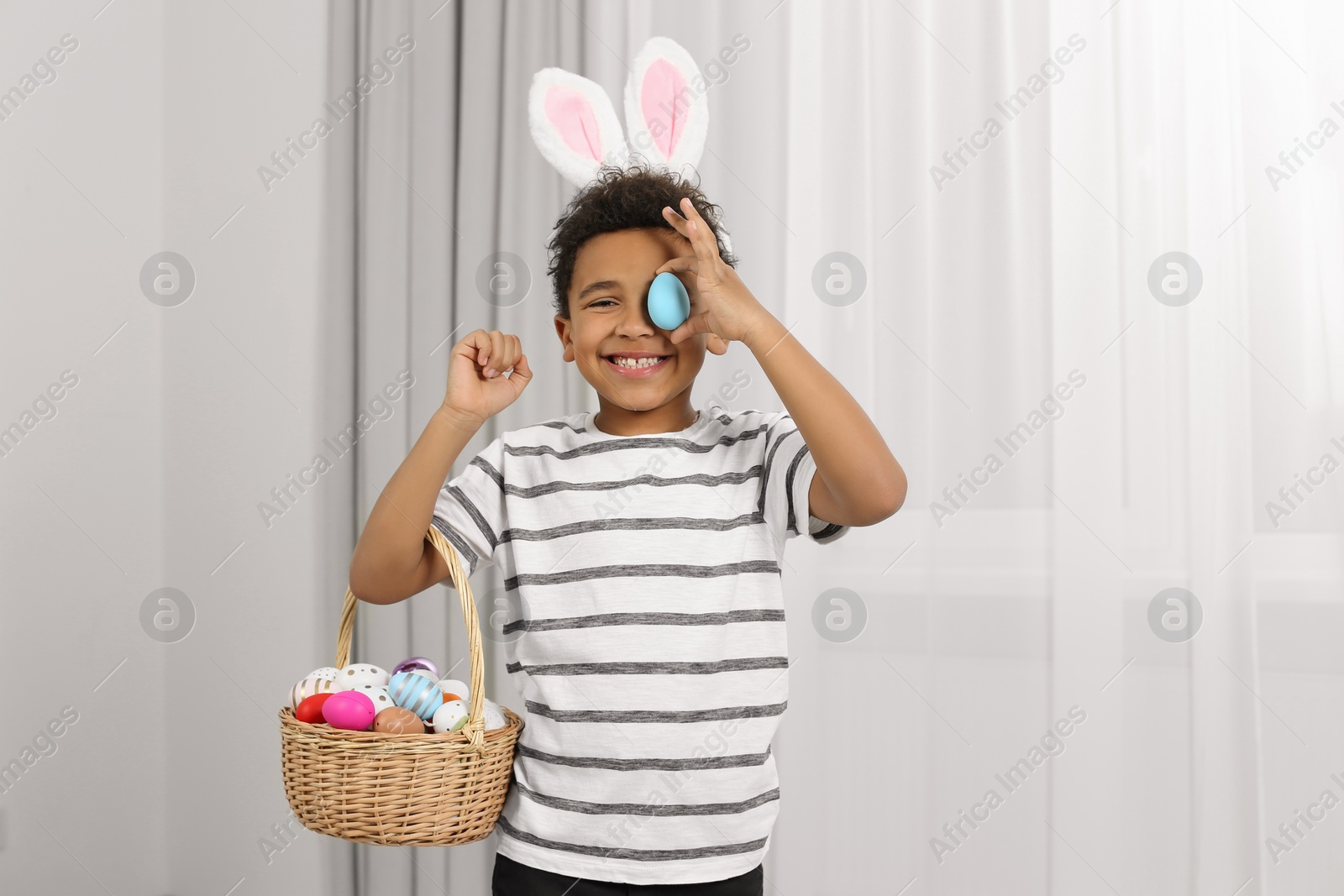 Photo of Cute African American boy in bunny ears headband covering eye with Easter egg indoors