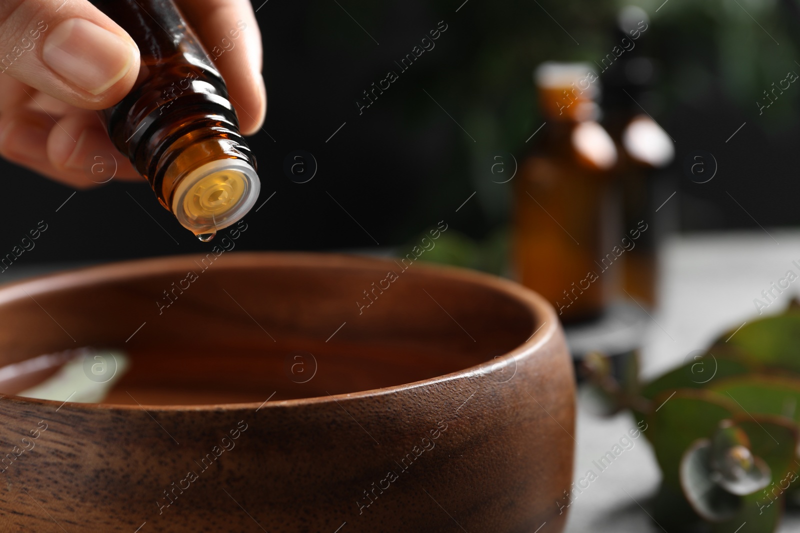 Photo of Woman dripping eucalyptus essential oil from bottle into bowl on blurred background, closeup. Space for text