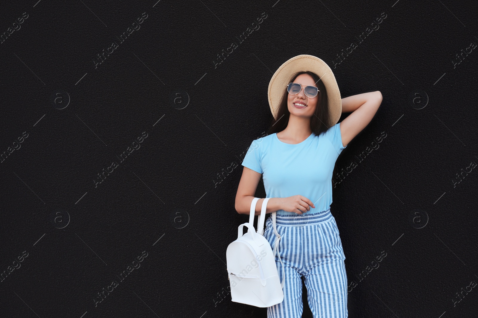 Photo of Beautiful young woman with stylish backpack and hat near black wall, space for text