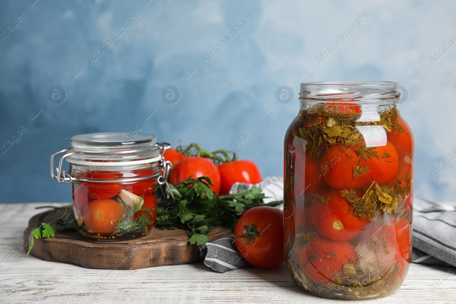 Photo of Pickled tomatoes in glass jars and products on white wooden table against blue background