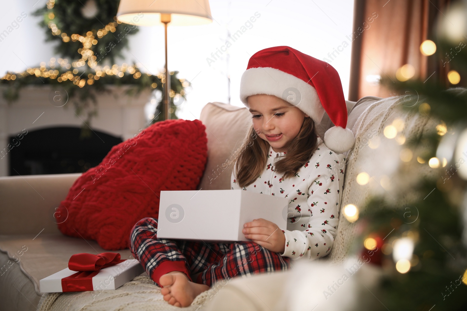 Photo of Cute little girl opening gift box on sofa in room decorated for Christmas