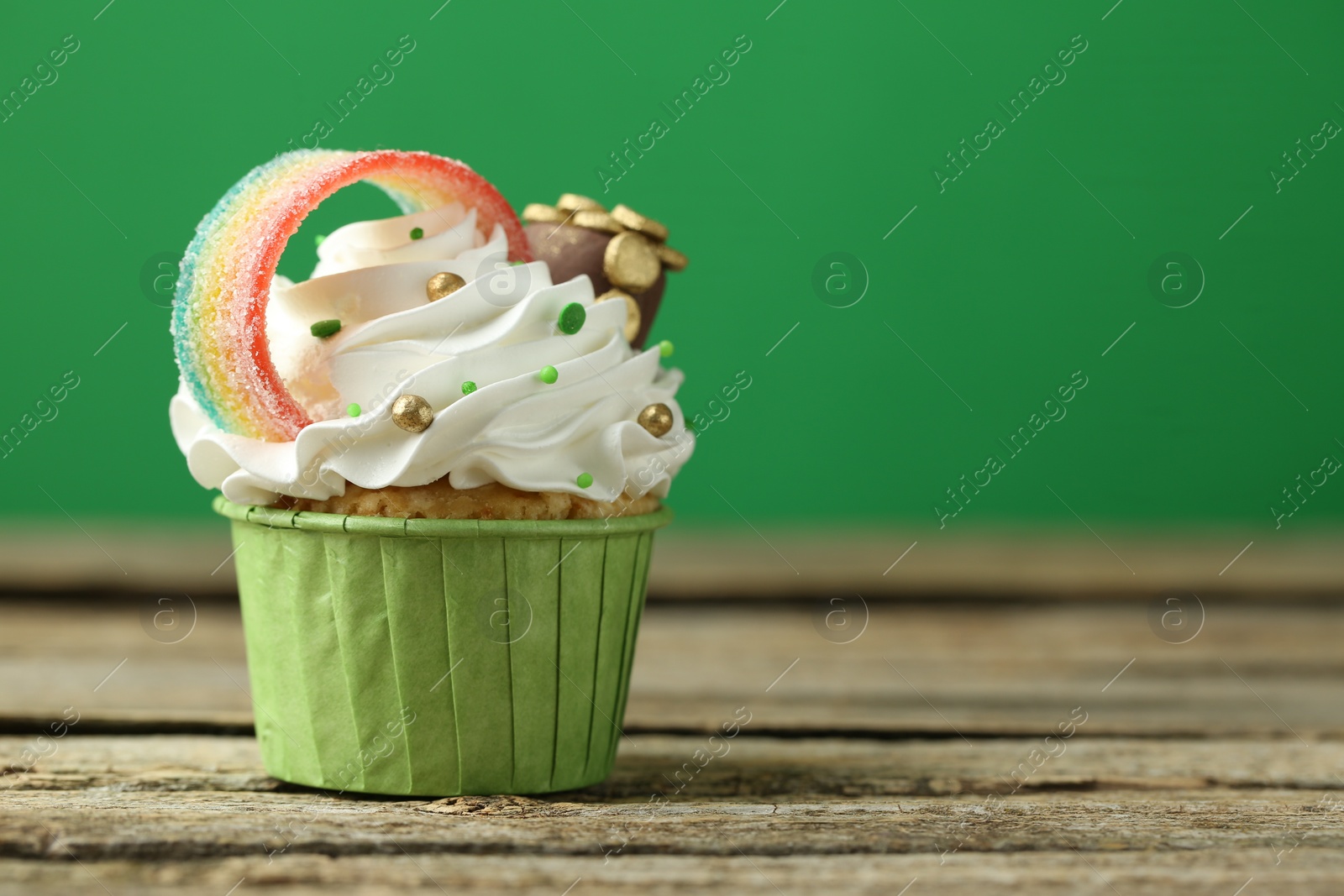 Photo of St. Patrick's day party. Tasty cupcake with sour rainbow belt and pot of gold toppers on wooden table, closeup. Space for text