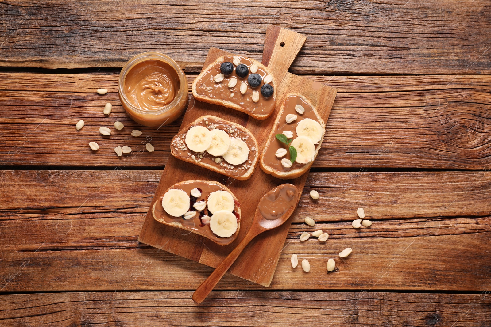 Photo of Toasts with tasty nut butter, banana slices, blueberries and peanuts on wooden table, flat lay