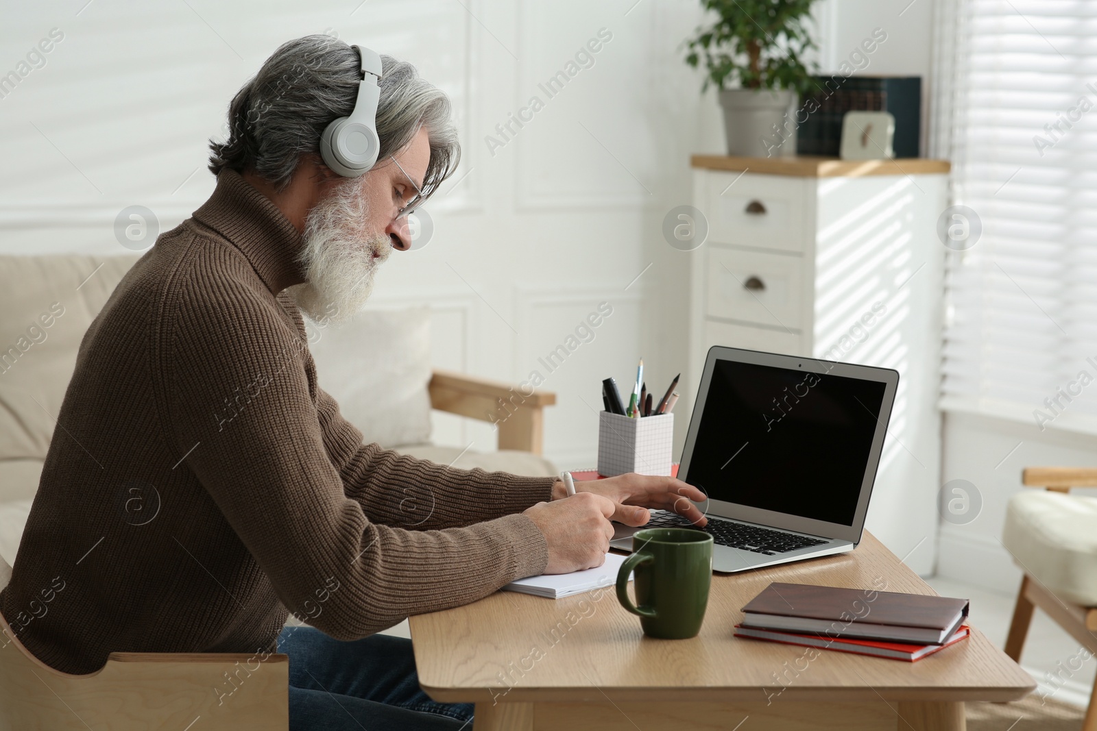 Photo of Middle aged man with laptop, notebook and headphones learning at table indoors