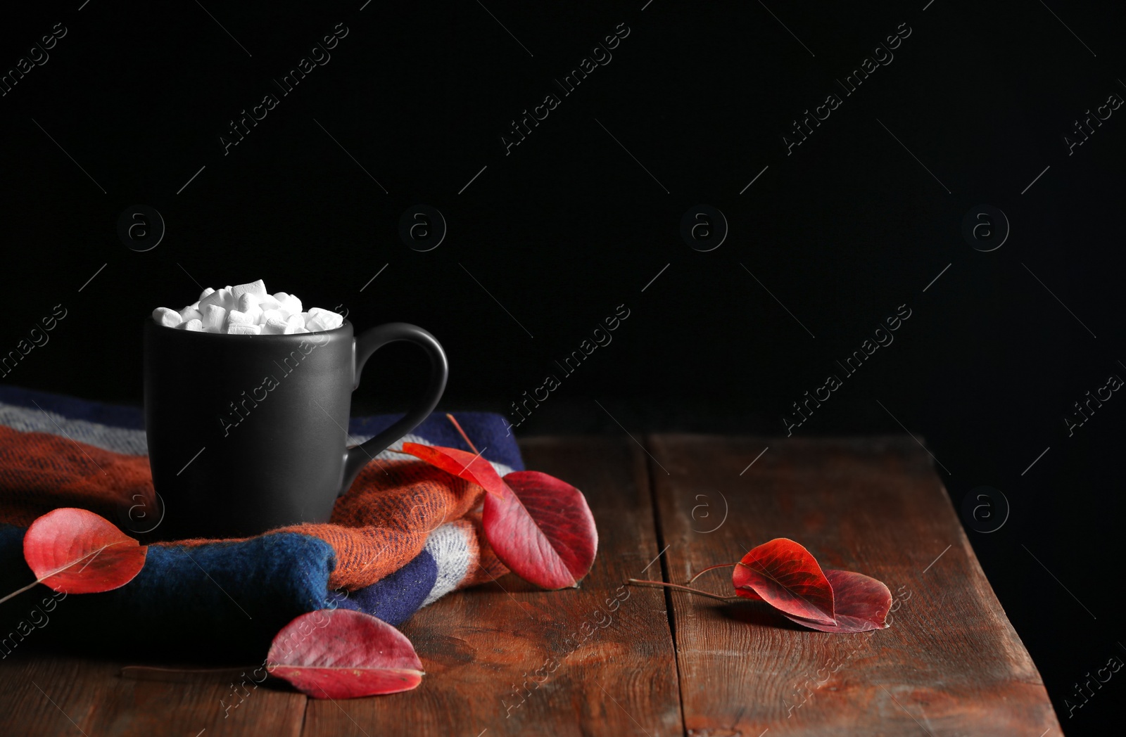 Photo of Cup of hot cozy drink with marshmallows, scarf and autumn leaves on table. Space for text