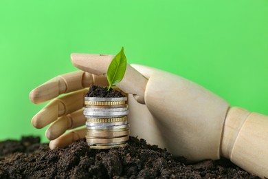 Stack of coins, green plant and wooden mannequin hand on soil against blurred background. Profit concept