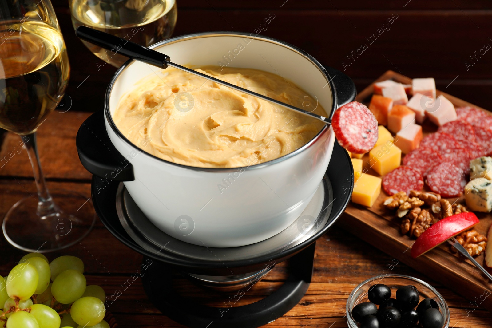 Photo of Fork with piece of sausage, melted cheese in fondue pot and other products on wooden table, closeup