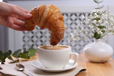 Tasty break. Woman dipping fresh croissant into cup with cappuccino at wooden table indoors, closeup